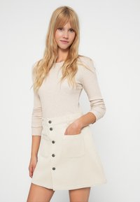 Beige ribbet langermet topp kombinert med et lyse beige kordfløyelskjørt med frontknapper og to sidelommer. Smooth tekstur.