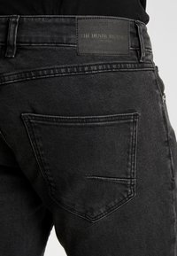 Jeans de denim negro con una etiqueta que dice "EL VIAJE DEL DENIM", un bolsillo trasero con detalles de costura y una tela suave y texturada.