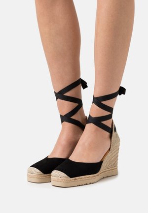 Tacchi a zeppa in suede nero con dettagli in juta intrecciata, fianchi aperti e nastri neri incrociati attorno alle caviglie. Design a punta rotonda.