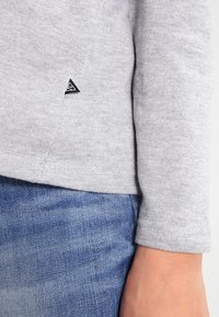 Felpa grigia realizzata in tessuto morbido, caratterizzata da un piccolo logo triangolare nero sul lato inferiore e maniche aderenti. Abbinata a jeans blu.