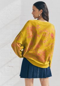Sweatshirt surdimensionné jaune avec des imprimés floraux et de paysage roses, associé à une jupe plissée en denim. Texture douce et coupe décontractée.