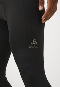 Schwarze Sportleggings aus elastischem Material mit einer glatten Oberfläche und präziser Nahtverarbeitung, versehen mit einem dezenten weißen Odlo-Logo am Oberschenkel.