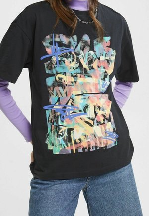 Osoba ubrana w czarny, oversize'owy T-shirt z kolorowym, abstrakcyjnym wzorem graffiti na lawendowych, długich rękawach i niebieskich dżinsach.