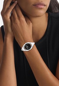 Montre-bracelet en silicone blanche avec un cadran circulaire noir, design minimaliste et accents argentés. Portée sur un poignet avec un haut noir texturé.