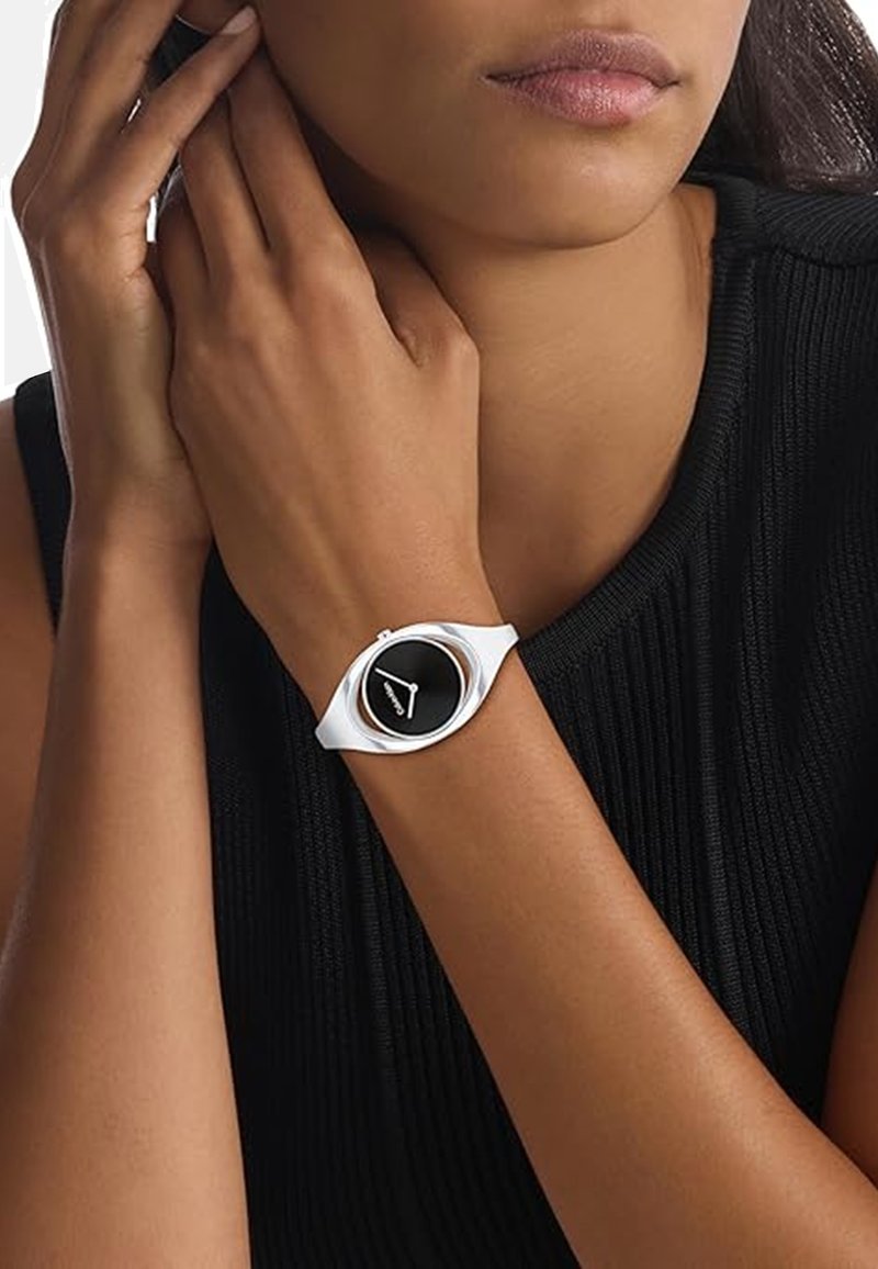 Montre-bracelet en silicone blanche avec un cadran circulaire noir, design minimaliste et accents argentés. Portée sur un poignet avec un haut noir texturé.