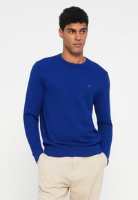 Kék textúrájú pulóver, kerek nyakkal és hosszú ujjakkal. Kis logó található a mellkasán, valamint bordázott mandzsetták és szegély. Párosítsd világos bézs nadrággal.