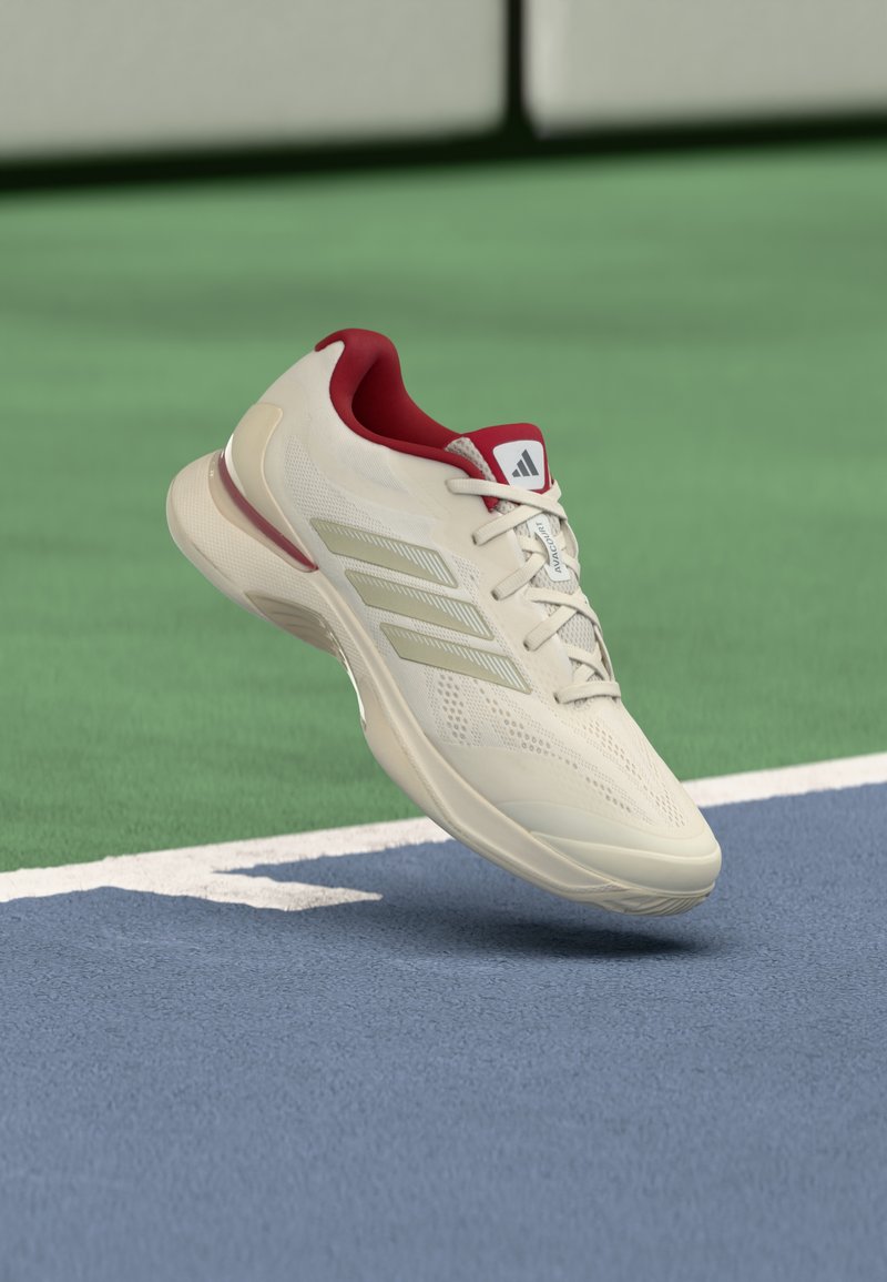 Kremowy but sportowy Adidas z czerwonym wnętrzem, unoszący się nad niebiesko-zieloną nawierzchnią kortu tenisowego w pobliżu białej linii granicznej.