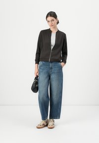 Svart bomberjacka med dragkedja fram, grå t-shirt under, vida blå jeans och beige sneakers. Modellen håller en svart handväska.