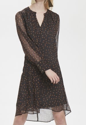 Woman wearing a black sheer long-sleeve dress with brown polka dots and buttoned cuffs, posing against a plain background.
