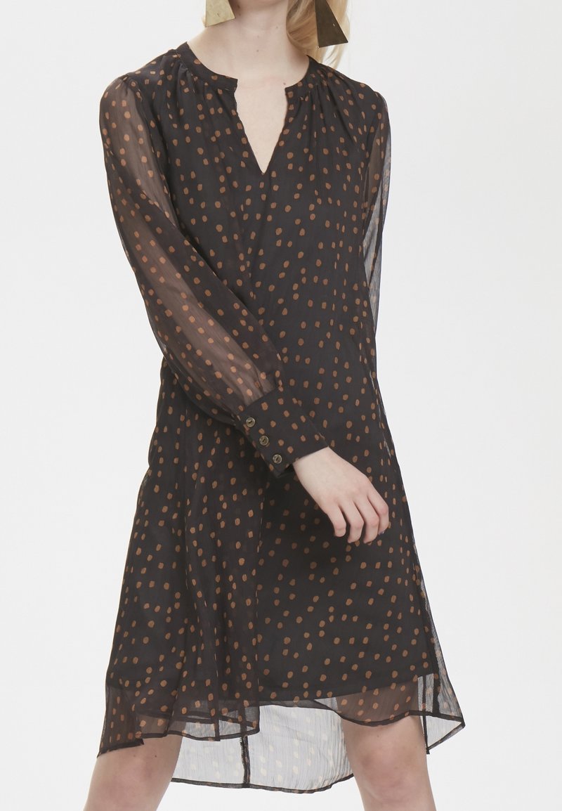 Femme portant une robe noire transparente à manches longues avec des pois marron et des poignets boutonnés, posant devant un fond uni.