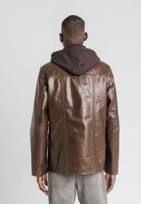 Bruine leren jas met een gladde textuur, rechte snit en zichtbare naden, gedragen over een donkere hoodie.