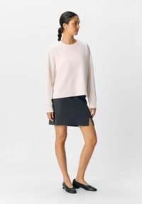 Helles rosa Sweatshirt mit langen Ärmeln, lockerer Schnitt, kombiniert mit einem dunkelgrauen Rock über dem Knie mit einem seitlichen Schlitz. Schwarze Ballerinas.