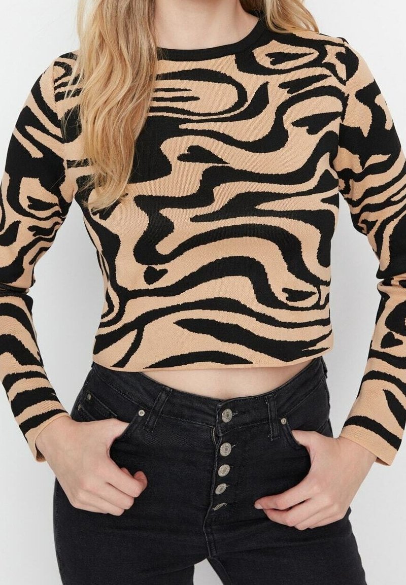Beige gestrickter, verkürzter Pullover mit schwarzem Wellenmuster, langen Ärmeln und rundem Ausschnitt, kombiniert mit hoch taillierten schwarzen Jeans.