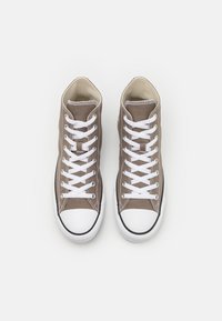 Wysokie trampki z brązowego płótna z białymi sznurówkami. Posiadają białe gumowe czubki i czarną lamówkę wzdłuż podeszwy. Gładka tekstura całości.