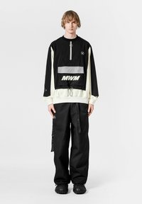 Suéter negro y crema con media cremallera, bolsillo frontal y detalles reflectantes. Combinado con pantalones negros texturizados y zapatos negros.