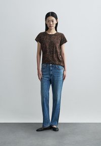 T-shirt com estampa de onça castanha, jeans azul de perna reta com desgaste mais claro, sapatos rasos pretos, posando contra um fundo neutro.