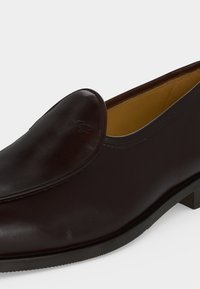 Σκούρο καφέ δερμάτινο παπούτσι slip-on με στρογγυλεμένη μύτη και διακριτικό ανάγλυφο λογότυπο στο πάνω μέρος, με χαμηλή μαύρη σόλα και ταμπά εσωτερική επένδυση.