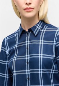 Woman with light blonde hair wearing a navy blue and white plaid button-up shirt with collar, shown from neck to mid-chest.