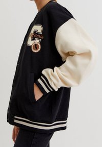 Černá varsity bunda s krémovými rukávy, na přední straně se velkým písmenem "C" a textem California. Pruhované manžety a lem, s kapsami.