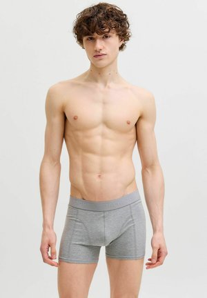 Ung mand med krøllet brunt hår iført tætsiddende grå boxershorts, stående foran en ensfarvet lys baggrund.