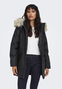 Vrouw in een zwarte winterjas met bontkraag, een wit shirt en donkere spijkerbroek, staand tegen een effen lichte achtergrond.