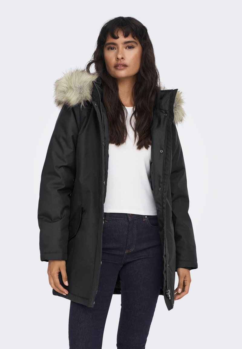 Vrouw in een zwarte winterjas met bontkraag, een wit shirt en donkere spijkerbroek, staand tegen een effen lichte achtergrond.