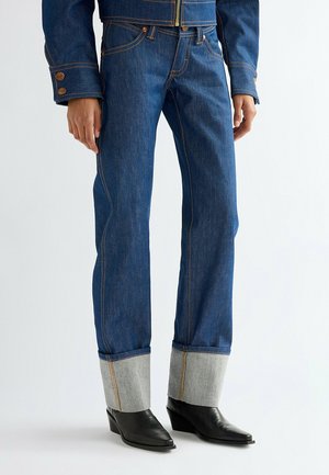 Person trägt dunkelblaue Jeans mit Umschlag und sichtbarer oranger Naht, kombiniert mit schwarzen spitzen Ankle Boots und einer passenden Jeansjacke.