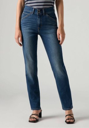 Jeans en denim bleu taille haute, jambe droite avec des patchs décolorés, cinq poches et fermeture à bouton en métal. Portés avec des talons noirs à lanières.