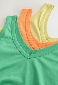 Pile de trois débardeurs en coton verts, orange et jaune, présentant des cols en V et des bords pliés, sur une surface neutre.