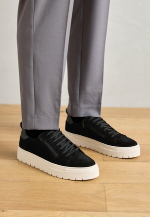Black suede sneakers with a textured finish, featuring a white rubber sole and decorative side zipper detail, worn with gray trousers.