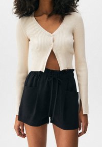 Žebrovaný krémový kardigan s výstřihem do V a dvěma knoflíky, kombinovaný s černými šortkami na stahovací šňůrku se bočními kapsami a volným střihem.
