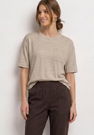 Femme souriante aux cheveux bruns en chignon portant une chemise beige à manches courtes avec poche poitrine et pantalon marron foncé à taille élastique, debout contre un mur blanc.