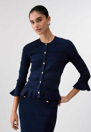 Frau trägt einen marineblauen, strukturierten, geknöpften Cardigan mit ausgestellten Bündchen und passendem Rock, steht mit einer Hand in der Hüfte vor weißem Hintergrund.