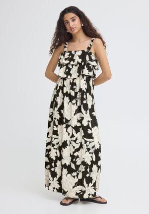 Femme aux cheveux longs et bouclés portant une robe maxi noire et blanche à fleurs avec un haut à volants et des sandales noires, debout les mains derrière le dos.