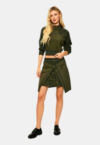 Donna bionda in maglione leopardato verde oliva e gonna coordinata con dettagli a strati, che indossa mocassini neri, in piedi contro uno sfondo semplice.