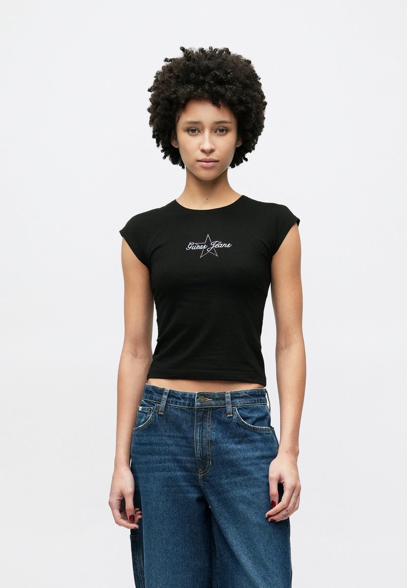 Zwarte katoenen cropped top met korte mouwen, met een zilveren sterprint en de tekst "Guess Jeans". Gecombineerd met blauwe jeans met wijde pijpen.
