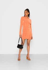 Vestido justo de cor pêssego, com fecho de botão na frente e mangas longas. O tecido texturizado cria um suave franzido. Combinado com uma pequena bolsa preta e saltos laranja.