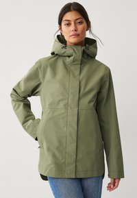 Chaqueta verde impermeable con cuello alto, capucha ajustable y dos bolsillos frontales. Textura suave y diseño de corte recto.