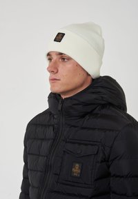 Giubbotto trapuntato nero con chiusura a zip e tasca con patta, abbinato a un berretto a coste di colore crema con un patch logo nero.