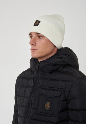 Giubbotto trapuntato nero con chiusura a zip e tasca con patta, abbinato a un berretto a coste di colore crema con un patch logo nero.