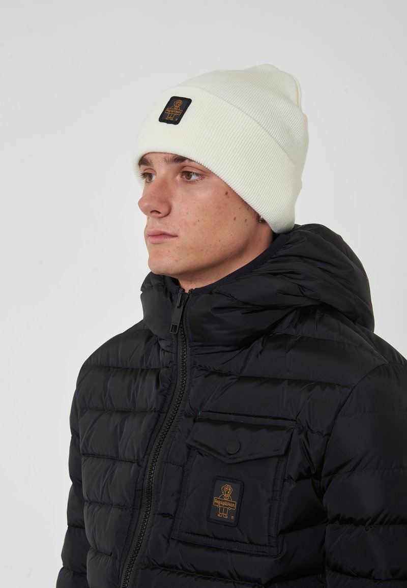 Giubbotto trapuntato nero con chiusura a zip e tasca con patta, abbinato a un berretto a coste di colore crema con un patch logo nero.