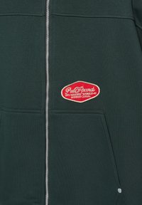 Mørkegrøn hættetrøje med lynlås, der har en central lynlås, to forlommer og et fremtrædende rødt broderet logo med tekst. Glat tekstur.