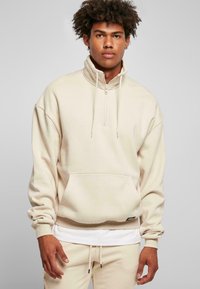 Pullover hettegenser i lys beige, laget av mykt stoff. Har høy krage, halv-glidelås foran og kengurulomme. Matchende bukser er synlige.