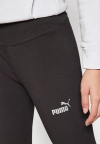 Juodos tamprės, pagamintos iš elastingos medžiagos, su lygiu juosmeniu ir baltu Puma logotipu, atspausdintu ant apatinės kojos.