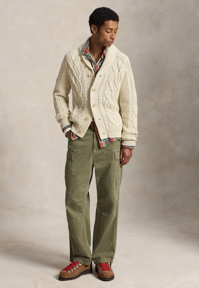 Άνδρας που φοράει μπεζ cardigan με πλεκτό σχέδιο, καρό πουκάμισο, ελαιί πράσινο παντελόνι cargo και καφέ ορειβατικά μποτάκια με κόκκινα κορδόνια, στέκεται με τα χέρια στις τσέπες.