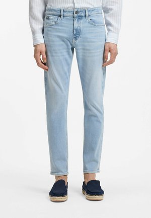 Hombre vistiendo jeans ajustados azul claro, camisa blanca con rayas y zapatos de esparto azul marino, de pie sobre un fondo blanco.