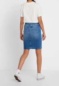 Denim kokerrok in middelblauw, voorzien van twee achterzakken en een rechte snit. Gecombineerd met een wit T-shirt met korte mouwen en witte sneakers.