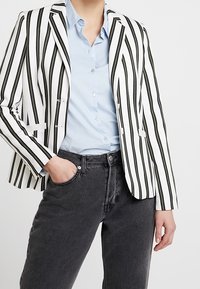 Pruhované sako v černé a bílé barvě, s na míru šitým střihem a úzkým střihem, nošené přes světle modrou košili a doplněné tmavými džínami z denimu.