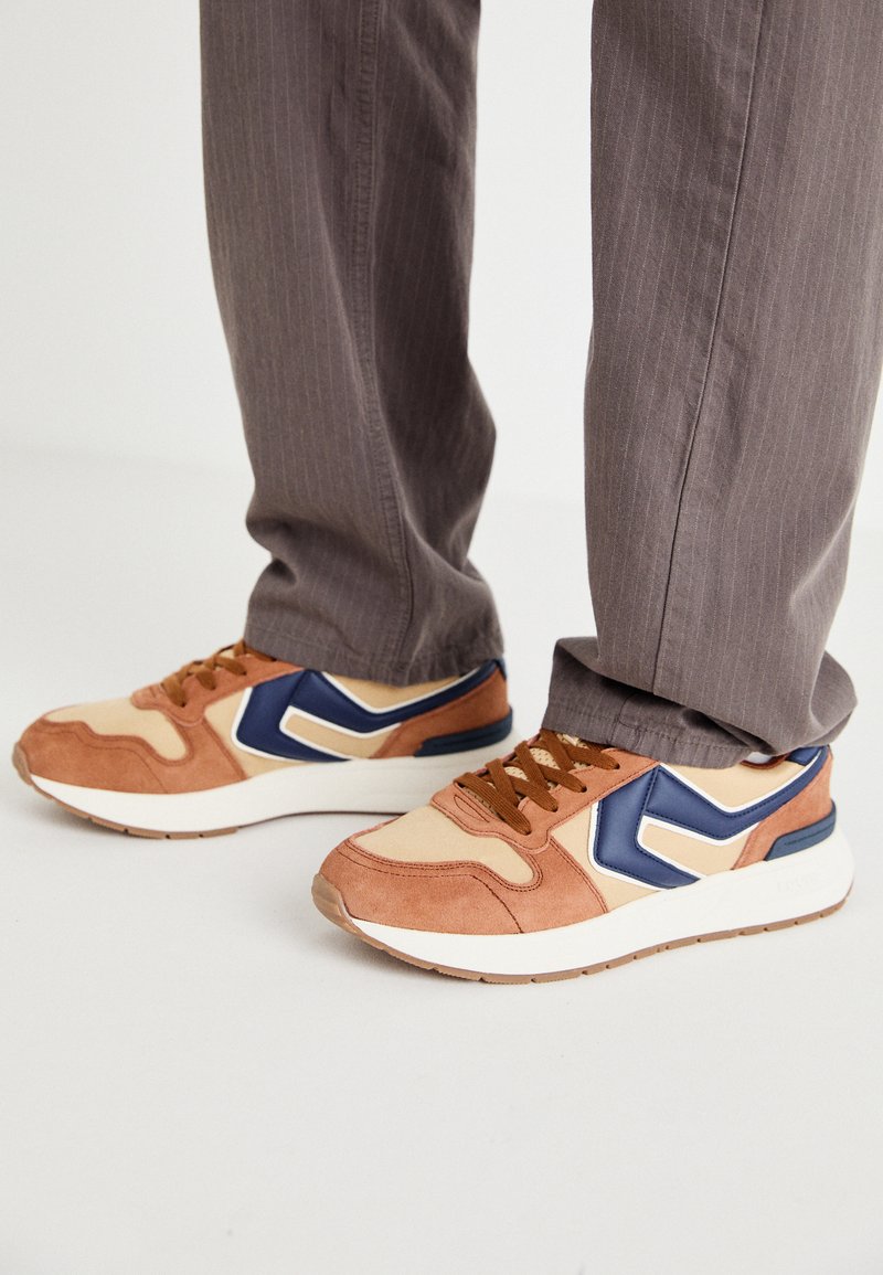 Sko med en blanding af tan ruskind og cremefarvet syntetisk materiale, med en blå accent og en struktureret sål; brune snørebånd; båret med grå stribede bukser.