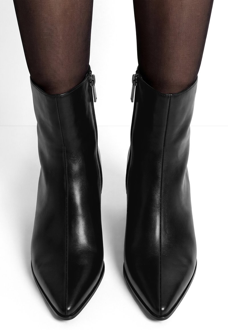 Paire de bottines en cuir noir à bout pointu portées avec des collants noirs transparents, vues de face sur fond blanc.